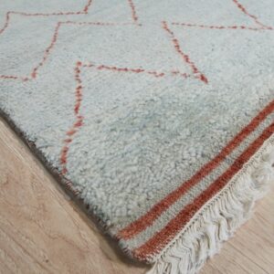 Shaggy off-white rug with a rust geometric pattern, striped border, and fringe on wood floor.