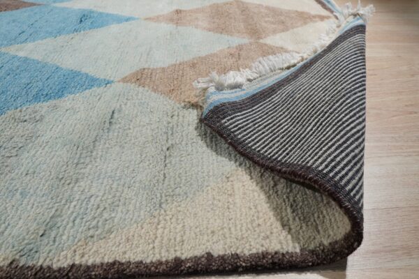 A rolled-back, shaggy rug with blue and beige diamonds rests on wood flooring, revealing a striped backing.