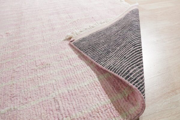 Pink high-pile rug showing horizontal stripes and a black and pink woven backing on light wood flooring.