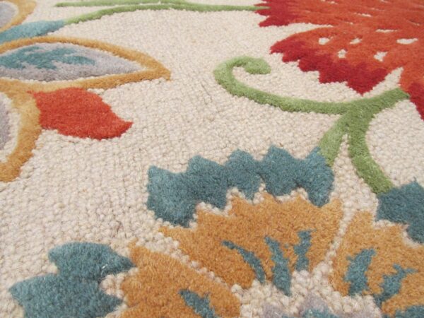 A textured rug close-up showing raised floral patterns in red, teal, gold, and green on a cream base.