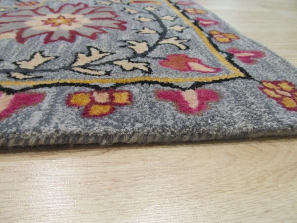 A close-up of a gray-blue floral rug with pink, yellow, and cream designs resting on light wood flooring.