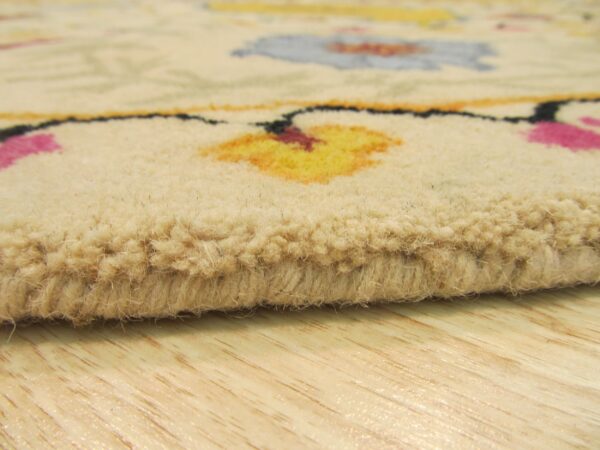 Close-up shows a thick beige rug pile with blurred yellow, pink, and blue floral motifs resting on light wooden flooring.