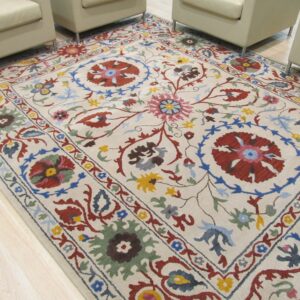 Medium-pile cream rug featuring a complex multicolored botanical design, partially framed by light armchairs on wood flooring.