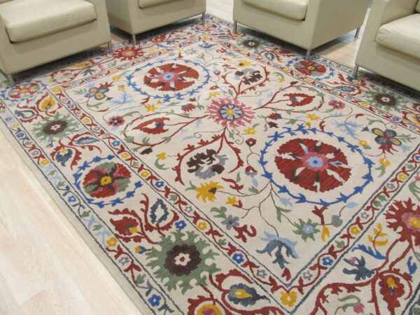 Medium-pile cream rug featuring a complex multicolored botanical design, partially framed by light armchairs on wood flooring.