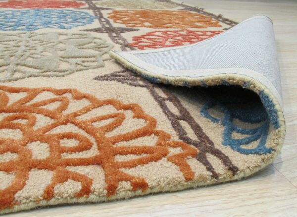 The rolled edge of a beige high-low pile rug displays blue, orange, and brown floral designs on wood flooring.
