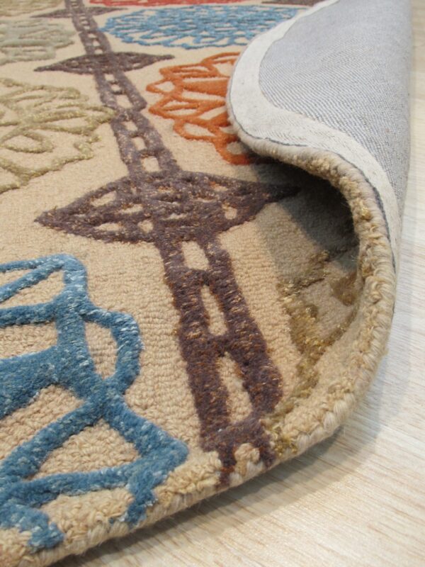 A textured beige rug with raised blue, orange, and brown patterns is curled, revealing the backing on a wood floor.