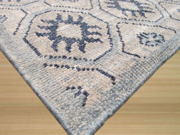 The corner of a textured blue and white rug with geometric patterns rests on light hardwood flooring.