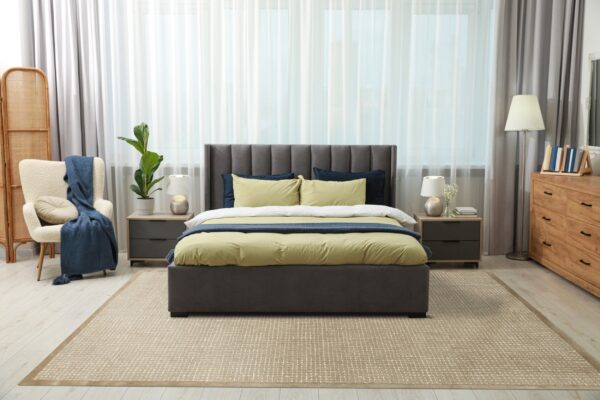 A textured tan rug with a woven grid pattern lies beneath a gray upholstered bed on light wood flooring.
