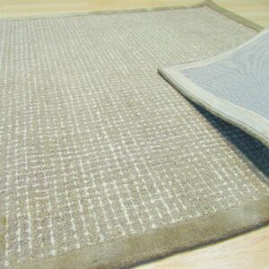 Tan rug with subtle textured geometric pattern lies on wood floor, showing the solid border and woven backing.