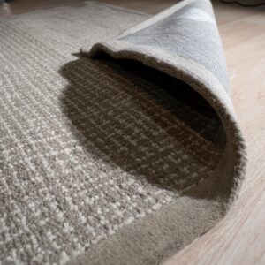 Textured beige and cream striped rug detail, showing the gray backing curled up on light wood flooring.