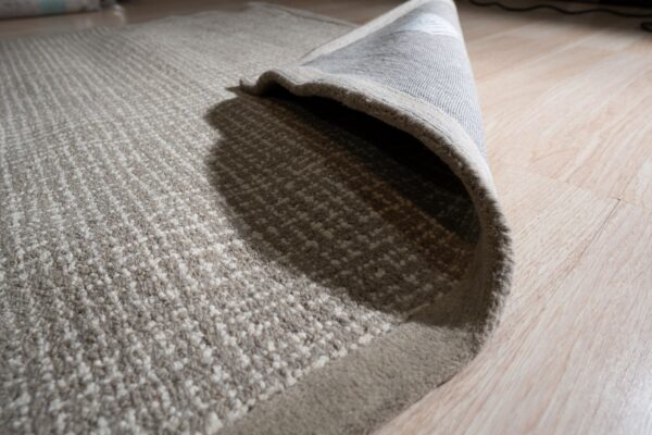 Textured beige and cream striped rug detail, showing the gray backing curled up on light wood flooring.