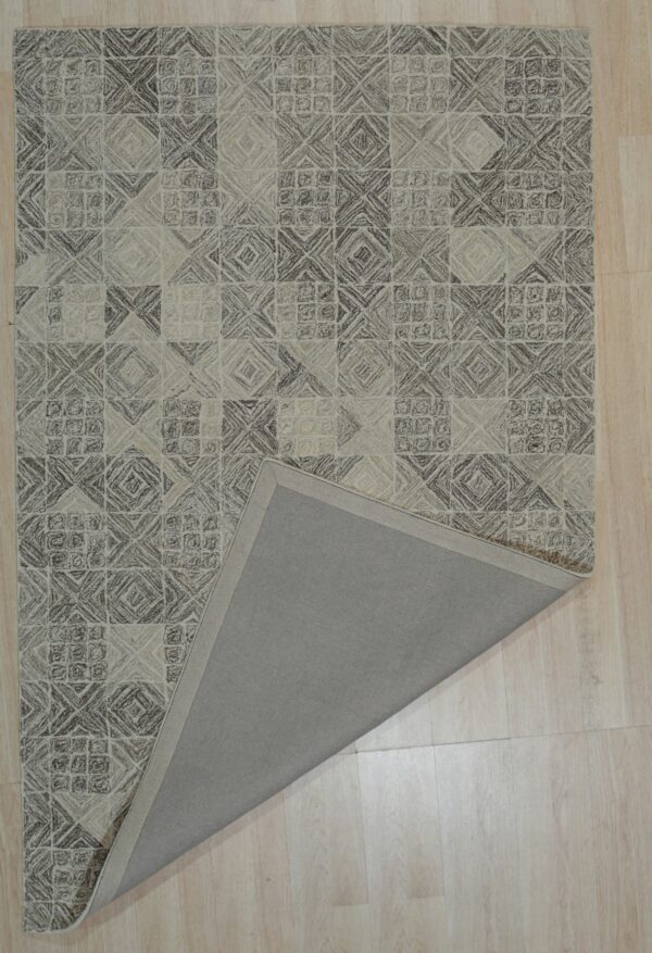 A beige and brown textured geometric rug lies on wood planks, folded back to show the plain gray backing.