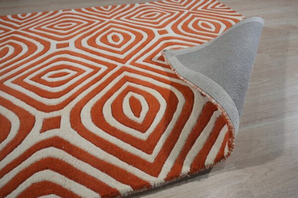 Close-up of a high-pile orange and cream geometric rug showing the woven gray backing on light flooring.