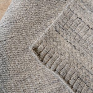 Close-up shows a light beige and gray mottled rug, folded over to reveal a shaggy diamond pattern on wood flooring.