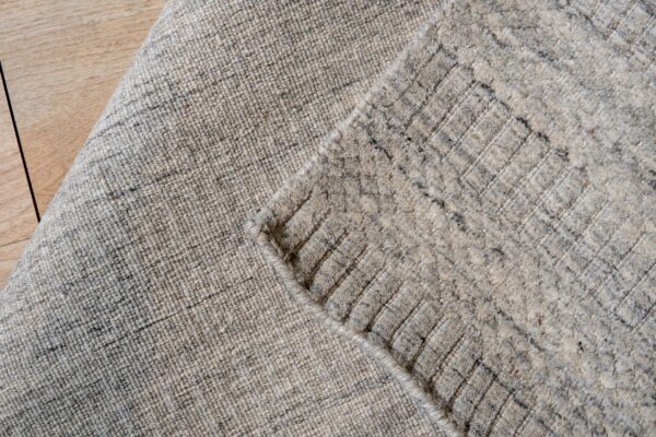 Close-up shows a light beige and gray mottled rug, folded over to reveal a shaggy diamond pattern on wood flooring.