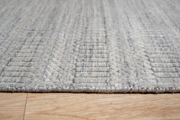 Close-up shows a heathered gray rug with a textured geometric pattern resting on light wood flooring.