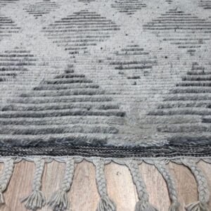 Textured light gray rug with dark geometric diamond patterns and braided fringe rests on wood plank flooring.