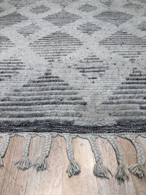 Textured light gray rug with dark geometric diamond patterns and braided fringe rests on wood plank flooring.