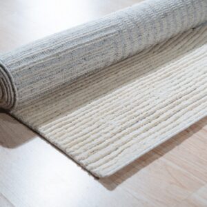 A ribbed cream and gray striped rug is partially unrolled onto light wood-look flooring.