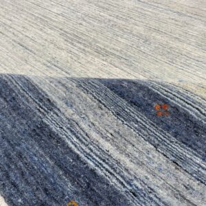 Textured rug surface partially rolled, showing light gray pile above dark blue and gray stripes with isolated orange patterns.