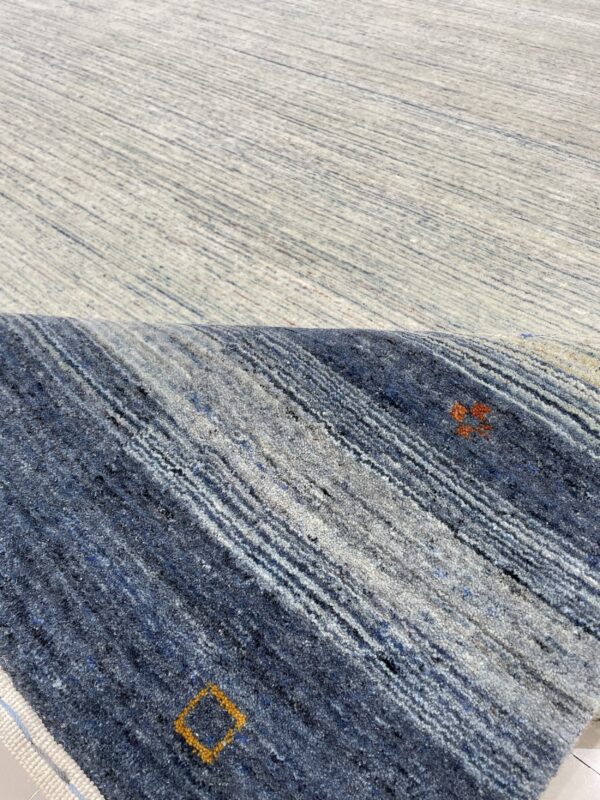 Textured rug surface partially rolled, showing light gray pile above dark blue and gray stripes with isolated orange patterns.