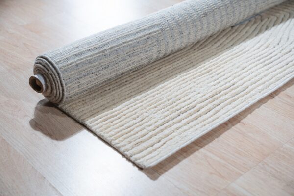 A cream and gray ribbed rug is partially unrolled on light wood plank flooring.