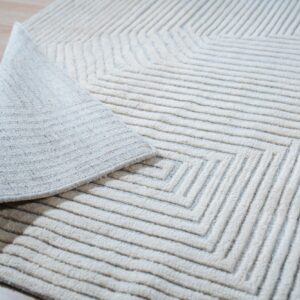 Off-white textured rug featuring deep grooved concentric lines, with a corner folded back on light wood flooring.