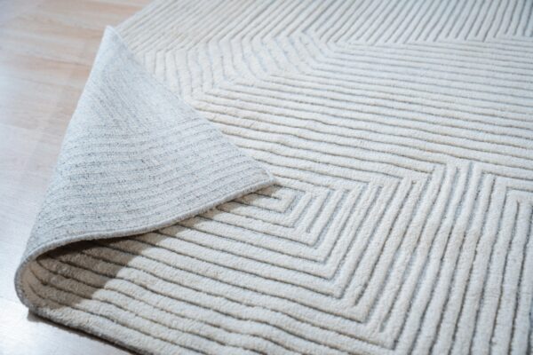 Off-white textured rug featuring deep grooved concentric lines, with a corner folded back on light wood flooring.
