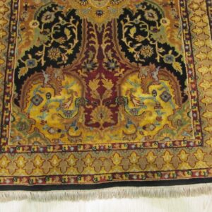 Gold, black, and red traditional rug with intricate floral patterns and a visible white fringe on light wood.