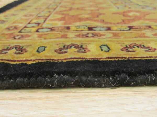The black, high-pile edge of a gold and rust patterned rug sits on light wood planks.