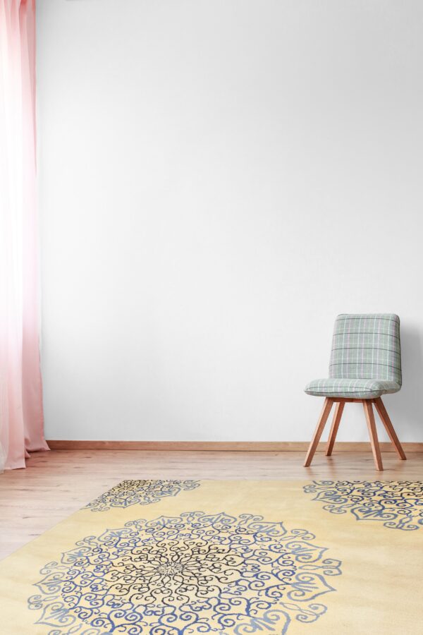 A light yellow rug with large blue and black medallion patterns is placed on a wood floor next to a checked chair.