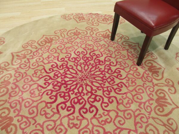 Round beige rug with a pink medallion pattern on wood floor, partially covered by a red chair.