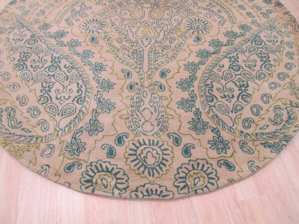 Round tan rug with dense teal and green paisley and floral patterns on a light wood floor.