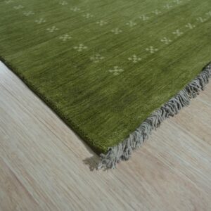 Close-up of a green pile rug with small beige crosses and gray fringe on light wood plank flooring.