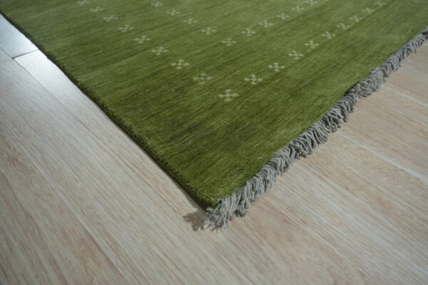 Close-up of a green pile rug with small beige crosses and gray fringe on light wood plank flooring.