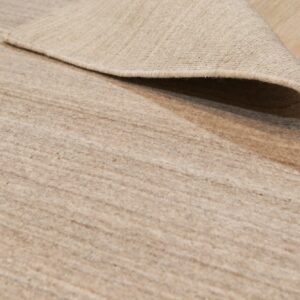 Low-pile tan rug with subtle striations, partially curled back to show the woven reverse side on light wood flooring.