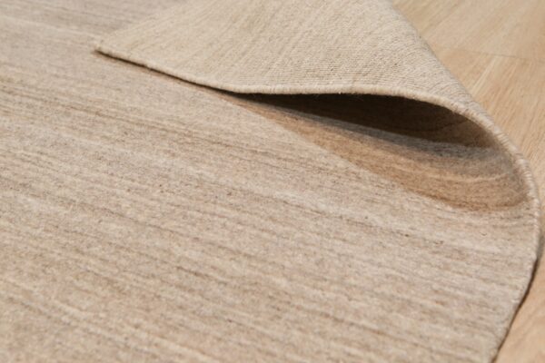 Low-pile tan rug with subtle striations, partially curled back to show the woven reverse side on light wood flooring.