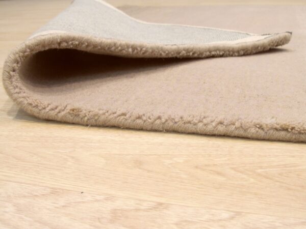 A beige rug with dense, low pile is folded over, showing the backing on light wood floor.