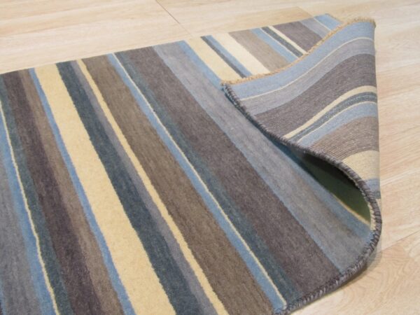 Multicolored striped rug in blue, gray, and tan is shown partially rolled on light wood flooring.