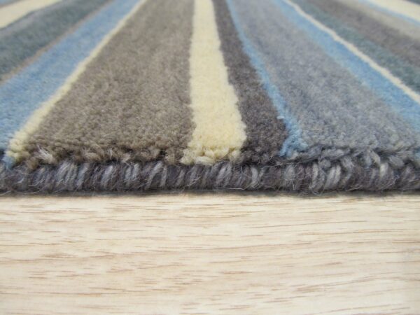 Close up of a striped rug edge with blue, gray, and cream medium pile on a light wood floor.