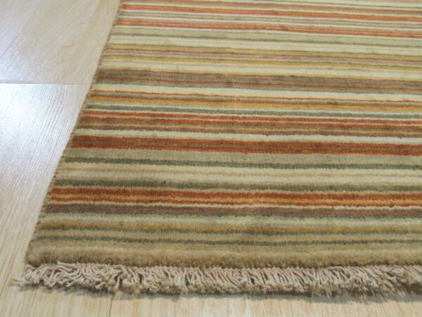 Earth-toned striped rug with beige, rust, and olive bands, showing a fringe detail on light wood flooring.