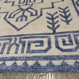 Angled view of a cream rug featuring textured blue geometric symbols and fringe over wood planks.