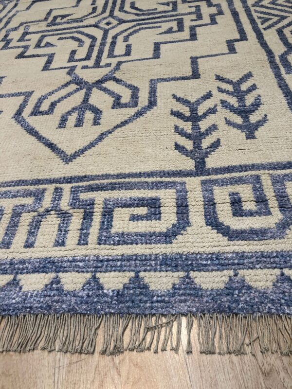 Angled view of a cream rug featuring textured blue geometric symbols and fringe over wood planks.