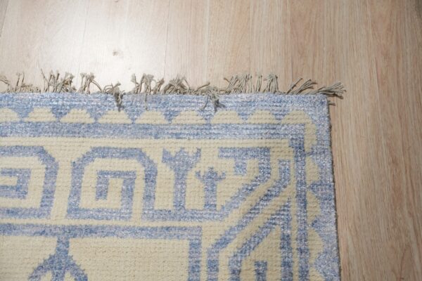 Light blue and cream rug with a geometric pattern, short fringe, and high pile on blonde wood flooring.