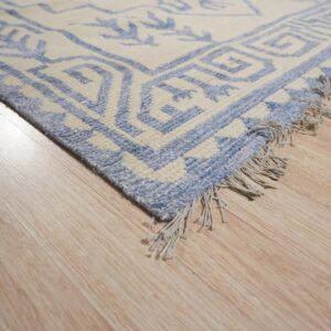 Textured cream and blue geometric rug corner with fringe resting on blonde wood planks.