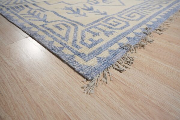 Textured cream and blue geometric rug corner with fringe resting on blonde wood planks.