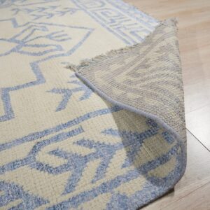 Cream rug with blue geometric patterns lifted to reveal backing, fringe, and thick pile on pale wood flooring.