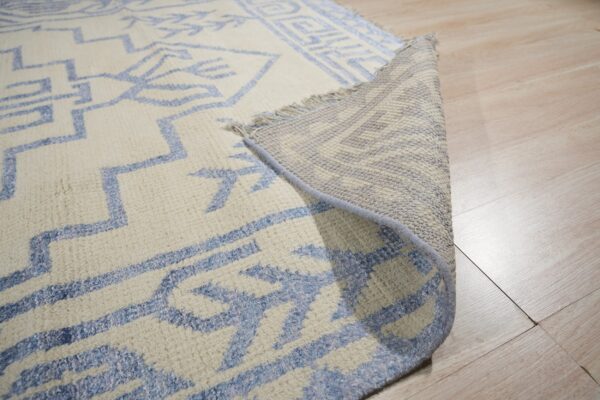 Cream rug with blue geometric patterns lifted to reveal backing, fringe, and thick pile on pale wood flooring.