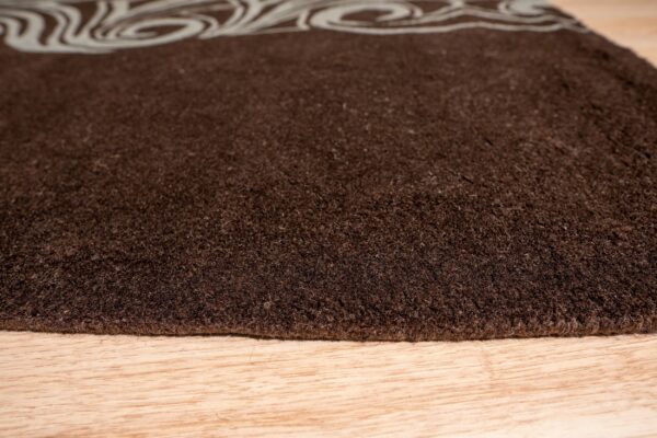 Dark brown high-pile rug with a light scroll pattern border lies on natural wood planks.