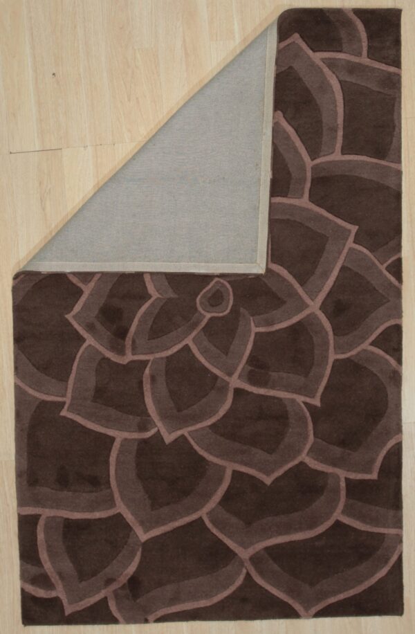 Dark brown carved rug with a repeating floral pattern, corner folded back revealing a gray underside on wood floor.